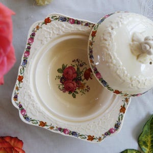 May include: A cream-colored ceramic serving dish with a matching lid, featuring a central floral bouquet of red roses and small colorful blossoms. The rim and lid are decorated with a delicate embossed floral pattern and a floral border.