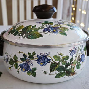 Könnte beinhalten: Ein weißer Emaille-Kochtopf mit passendem Deckel, verziert mit einem botanischen Muster aus blauen Akelei-Blüten und grünen Blättern. Der Topf hat zwei dunkelbraune Seitengriffe und einen passenden dunkelbraunen Knauf am Deckel sowie einen silbernen Metallrand.