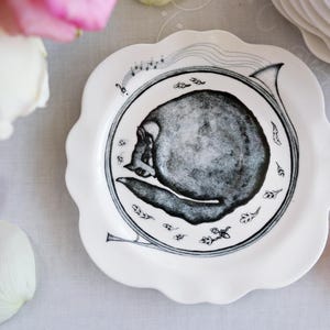 May include: White ceramic plate with a scalloped edge, featuring a black and gray illustration of a skull within a musical horn design. The plate is surrounded by pink and white rose petals.