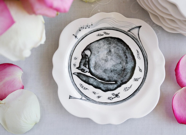 May include: White ceramic plate with a scalloped edge, featuring a black and gray illustration of a skull within a musical horn design. The plate is surrounded by pink and white rose petals.