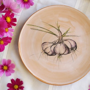 Puede incluir: Un plato de cerámica con un diseño pintado a mano de un bulbo de ajo con tallos verdes. El plato es de color beige claro.