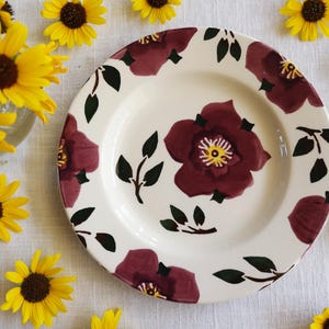 May include: A white ceramic plate with a burgundy floral pattern and green leaves. The plate is surrounded by yellow sunflowers. The floral design features large burgundy flowers with yellow centers, creating a rustic, charming aesthetic.