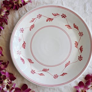 May include: White ceramic dinner plate with a pink floral design around the rim. The plate has a slightly raised edge.