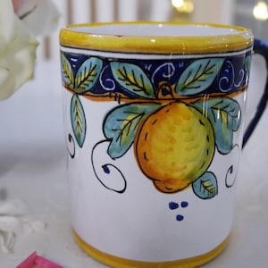 May include: White ceramic mug with a hand-painted design featuring a yellow lemon, green leaves, and blue accents. The mug has a yellow rim and handle, perfect for enjoying your favorite beverage. The mug is approximately 4 inches tall.