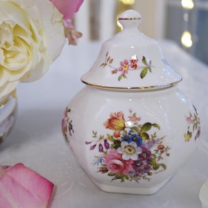 以下が含まれることがあります： ピンク、オレンジ、青、紫の色合いの花束のデザインが施された、蓋付きの白い磁器の壺。 壺には金のトリムと蓋に装飾的なつまみが付いています。 壺はおそらく装飾用です。