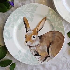 Può includere: Un piatto da portata in ceramica bianca con un motivo damascato verde chiaro e bianco. Un coniglio marrone con pelliccia bianca è dipinto sul piatto.