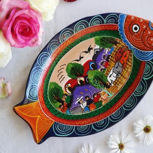 Puede incluir: Una fuente de cerámica pintada a mano con forma de pez. La fuente presenta un diseño vibrante con una cabeza roja, una cola naranja y una escena central con figuras, árboles y pájaros. El borde es azul marino con un patrón decorativo.