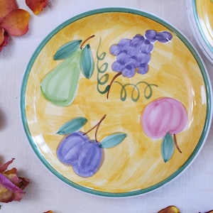 May include: A round, hand-painted ceramic plate with a yellow center and a green border. The plate features illustrations of a green pear, purple grapes, a pink peach, and purple plums. The design is in a whimsical, fruit-themed style.