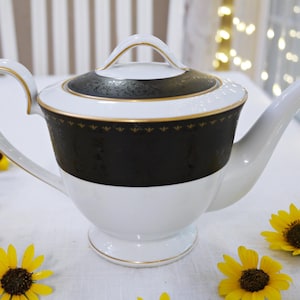 Może przedstawiać: Biało-czarny porcelanowy imbryk ze złotymi zdobieniami. Imbryk ma zakrzywiony uchwyt i dzióbek. Pokrywka jest czarna z dekoracyjnym wzorem. Żółte słoneczniki są rozrzucone wokół imbryka na białej powierzchni.