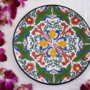 Puede incluir: Un plato decorativo de cerámica con borde negro. El plato presenta un diseño floral simétrico en rojo, amarillo, azul y verde sobre fondo blanco. El diseño incluye flores, hojas y patrones ornamentales.