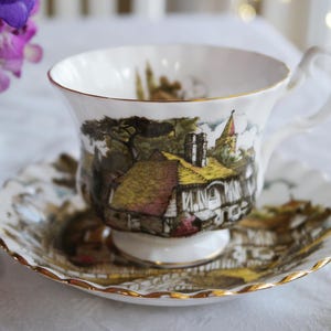 May include: A white teacup and saucer with a gold rim. The teacup and saucer feature a colorful illustration of a cottage with a thatched roof and a church steeple in the background.
