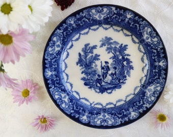 May include: A blue and white porcelain plate with a decorative border and a central image of a couple in a landscape setting.