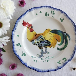 May include: White ceramic plate with a scalloped edge and a blue border. The plate features a colorful illustration of a rooster with yellow, blue, and green feathers. Green shamrocks are scattered around the plate's edge, adding a touch of whimsy.