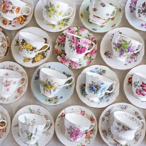 May include: An assortment of vintage teacups and saucers, each with unique floral patterns and gold trim. The cups and saucers are arranged on a neutral surface, showcasing a variety of colors including pink, yellow, blue, and purple.