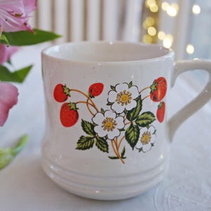 May include: Cream-colored ceramic mug featuring a strawberry and flower design. The mug has a curved handle and a slightly textured surface. The strawberries are red, and the flowers are white with yellow centers.