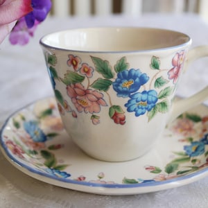 Puede incluir: Una taza de té y un platillo blancos con un patrón floral. El patrón presenta flores rosas, azules y verdes sobre un fondo blanco. La taza tiene un asa y el platillo tiene un borde festoneado.