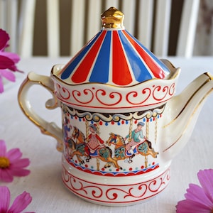 Op de afbeelding: Een witte keramische theepot met een carrouselontwerp. Het deksel is rood en blauw gestreept, met een gouden knop. De body heeft carrouselpaarden en -ruiters, met gouden randen en een rood-witte decoratieve rand.