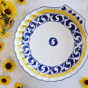 May include: A large, white ceramic serving platter with a scalloped edge. The platter features a yellow and blue decorative border with a repeating swirl design. A bouquet of yellow sunflowers is in the background.