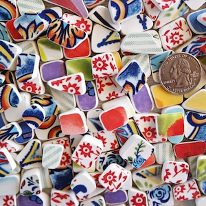 Könnte beinhalten: Eine Sammlung bunter Keramikfliesen mit verschiedenen Mustern, darunter florale Designs, geometrische Formen und abstrakte Kunst. Die Fliesen sind in einem zufälligen Muster angeordnet und überwiegend weiß mit Akzenten in Rot, Blau, Grün und Gelb. Eine deutsche 20-Cent-Münze ist zur Größenbestimmung enthalten.