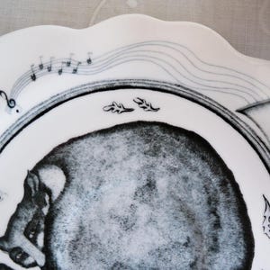 May include: White ceramic plate with a scalloped edge, featuring a black and gray illustration of a musical instrument and a stylized animal. Musical notes and decorative leaves are also included in the design.