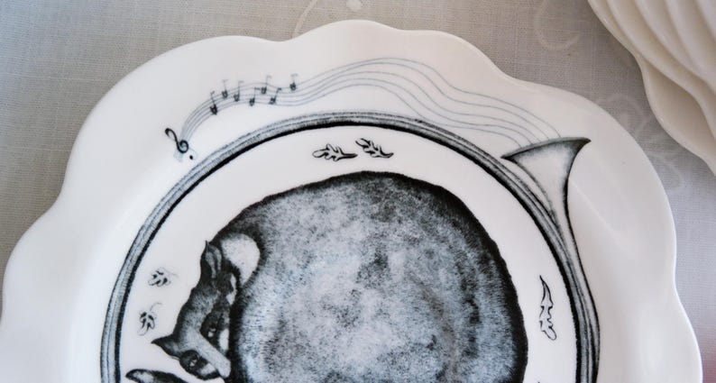 May include: White ceramic plate with a scalloped edge, featuring a black and gray illustration of a musical instrument and a stylized animal. Musical notes and decorative leaves are also included in the design.