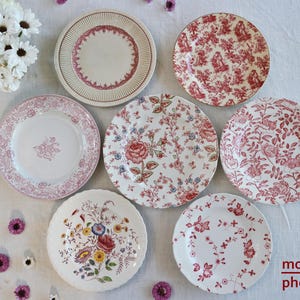 May include: A collection of vintage ceramic plates with various floral and patterned designs in shades of red, pink, and white. The plates are arranged on a white surface, showcasing their different styles and sizes. Some plates feature intricate floral patterns, while others have simpler, more geometric designs.