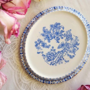 May include: An oval ceramic platter with a cream-colored base and a blue floral pattern in the center. The rim features a detailed blue and white geometric border with a thin gold-colored edge. The design includes stylized blossoms.