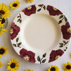 May include: A white ceramic plate with a maroon floral pattern around the rim. The flowers have yellow centres and green leaves. Sunflowers are scattered around the plate.