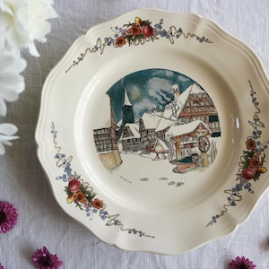 Puede incluir: Plato decorativo color crema con borde festoneado. El plato presenta una escena de pueblo invernal en el centro y detalles florales y de cinta alrededor del borde. La escena del pueblo incluye edificios cubiertos de nieve y una iglesia.