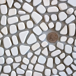 Puede incluir: Una imagen de primer plano de un patrón de mosaico de azulejos blancos. Los azulejos son de varias formas y tamaños, que se asemejan a guijarros. Una moneda de euro española de 1 euro se coloca en el centro de la imagen para la escala.