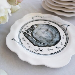 May include: White ceramic plate with a scalloped edge, featuring a black and gray illustration of a snail. The plate has a detailed design with small leaf and line art accents. A stack of similar plates is in the background.