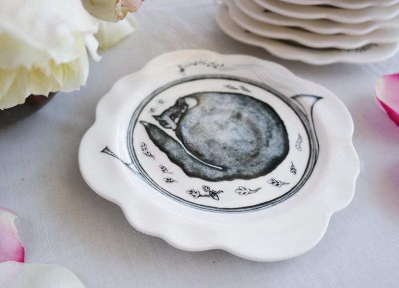 May include: White ceramic plate with a scalloped edge, featuring a black and gray illustration of a snail. The plate has a detailed design with small leaf and line art accents. A stack of similar plates is in the background.