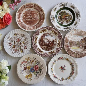 May include: A collection of vintage decorative plates with various designs. The plates feature images of ships, landscapes, and floral patterns in shades of brown, green, and white. The plates are arranged on a white surface.