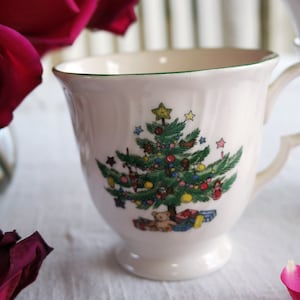 Puede incluir: Taza de té de cerámica blanca con un borde verde y un diseño de árbol de Navidad. El árbol está decorado con adornos, estrellas y un oso de peluche.