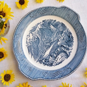 Puede incluir: Un plato decorativo azul y blanco con una escena de paisaje en el centro. El plato tiene un borde festoneado y está rodeado de girasoles amarillos. El borde del plato presenta un diseño texturizado.