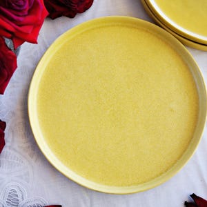May include: A round, solid yellow dinner plate with a smooth, matte finish and a slightly raised rim. The plate is positioned centrally, accompanied by deep red roses and additional yellow plates stacked in the upper right corner.