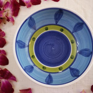 Könnte beinhalten: Eine handbemalte Keramikschale mit blauem und weißem Design. Die Schale hat eine zentrale dunkelblaue Spirale, umgeben von einem grün-gelben Ring und blauen blattförmigen Akzenten. Die Schale hat einen weißen Rand und wird wahrscheinlich zum Servieren von Speisen verwendet.