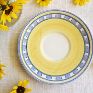 May include: A ceramic plate with a yellow and blue design. The plate has a white centre and a blue border. Yellow sunflowers are arranged around the plate and in a glass vase.