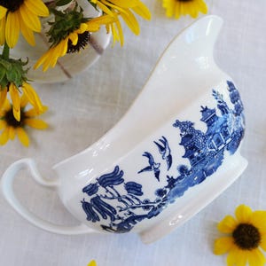 May include: White porcelain gravy boat with a blue Willow pattern design. The gravy boat has a curved handle and a spout. Yellow sunflowers are scattered around the gravy boat.
