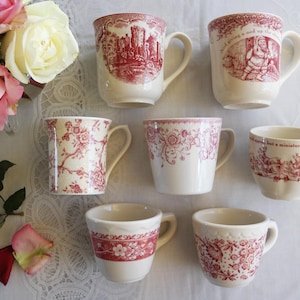 May include: A collection of cream-colored ceramic mugs with red designs. The mugs feature various patterns, including floral, castle, and pastoral scenes. Some mugs have text. A bouquet of pink and white roses is in the background.