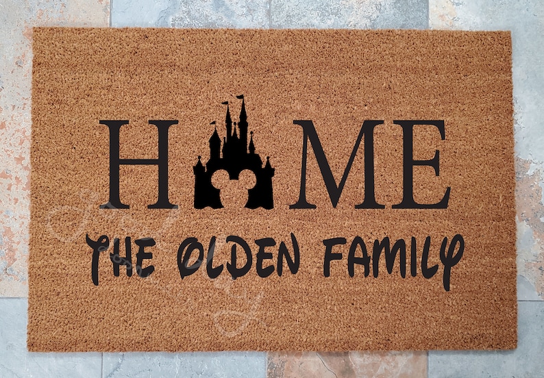 K&ouml;nnte beinhalten: Eine braune Kokosmatte mit dem Text "HOME" in schwarzen Buchstaben, einer schwarzen Silhouette des Disney-Schlosses mit Mickey-Maus-Ohren in der Mitte des "O" und dem Text "THE OLDEN FAMILY" in schwarzen Buchstaben.