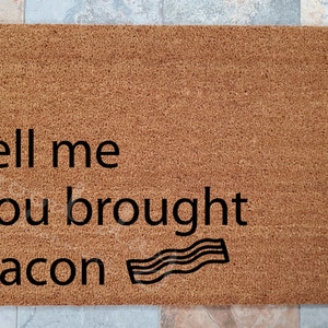 May include: A rectangular doormat made of brown coir fiber. The mat features the phrase "tell me you brought bacon" in black text, with a stylized bacon strip graphic.