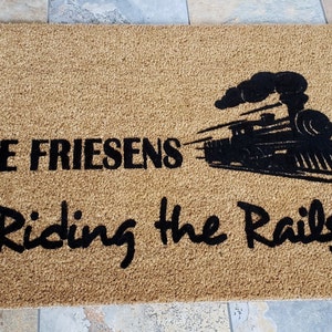 May include: A brown coir doormat with the text "The Friesens Riding the Rails" in black lettering. A black silhouette of a train with smoke is in the center of the mat.
