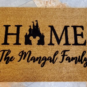 K&ouml;nnte beinhalten: Eine braune Kokosmatte mit dem Wort "HOME" in schwarzen Buchstaben, einer schwarzen Silhouette eines Schlosses mit Mickey-Maus-Ohren und dem Text "The Mangal Family" in schwarzer Kursivschrift.