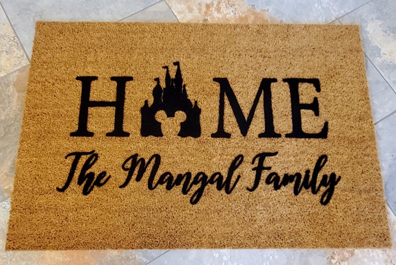K&ouml;nnte beinhalten: Eine braune Kokosmatte mit dem Wort "HOME" in schwarzen Buchstaben, einer schwarzen Silhouette eines Schlosses mit Mickey-Maus-Ohren und dem Text "The Mangal Family" in schwarzer Kursivschrift.