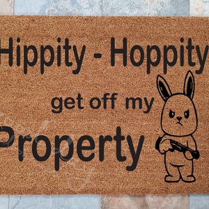 May include: A rectangular doormat made of brown coir fiber. The mat features the text "Hippity - Hoppity get off my Property" in black, along with a cartoon rabbit holding a rifle.