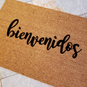 以下が含まれることがあります： 黒い筆記体で「bienvenidos」と書かれた茶色のココナッツ繊維のドアマットです。