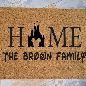 K&ouml;nnte beinhalten: Eine braune Kokosmatte mit dem Text "HOME" in Schwarz, die eine schwarze Silhouette des Disney-Schlosses mit Mickey-Maus-Ohren zeigt. Der Text "THE BROWN FAMILY" ist ebenfalls in Schwarz unterhalb des Schlosses.