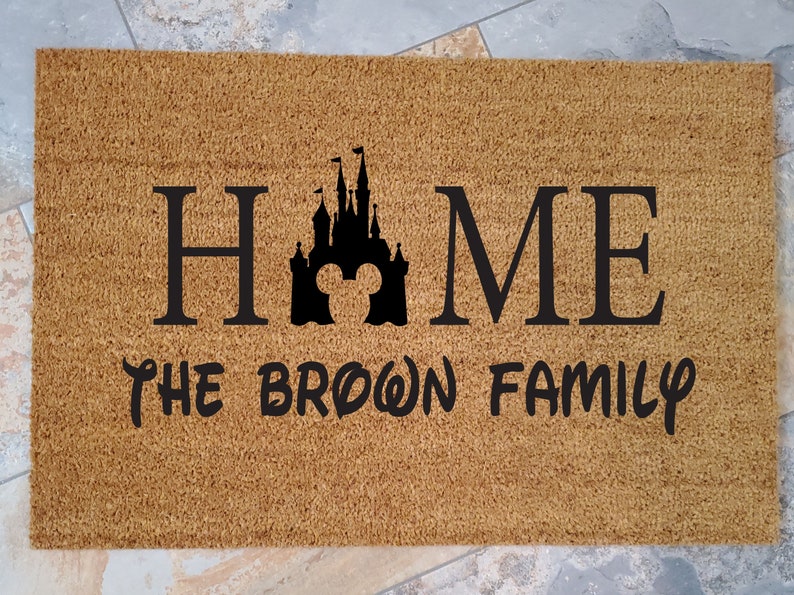 K&ouml;nnte beinhalten: Eine braune Kokosmatte mit dem Text "HOME" in Schwarz, die eine schwarze Silhouette des Disney-Schlosses mit Mickey-Maus-Ohren zeigt. Der Text "THE BROWN FAMILY" ist ebenfalls in Schwarz unterhalb des Schlosses.