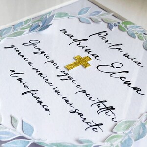 Puede incluir: Una tarjeta blanca con una cruz dorada y una corona de laurel acuarela azul y verde. La tarjeta dice "Per la mia madrina Elena Grazie per oggi e per tutti i giorni a venire in cui sarai al mio fianco."